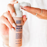 Hands holding Noosa Basics Foaming Face Wash and dispensing a mound of white cleansing foam onto the fingertips.