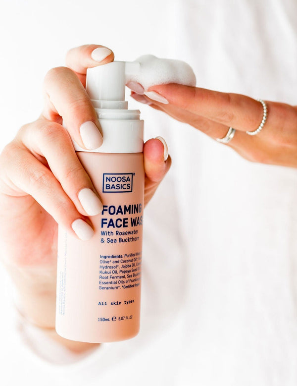 Hands holding Noosa Basics Foaming Face Wash and dispensing a mound of white cleansing foam onto the fingertips.