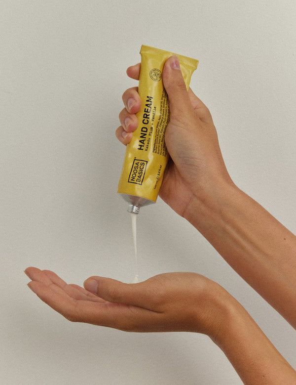 Hands squeezing a thin stream of white Noosa Basics Kakadu Plum and Vanilla Hand Cream from a yellow tube into an open palm.
