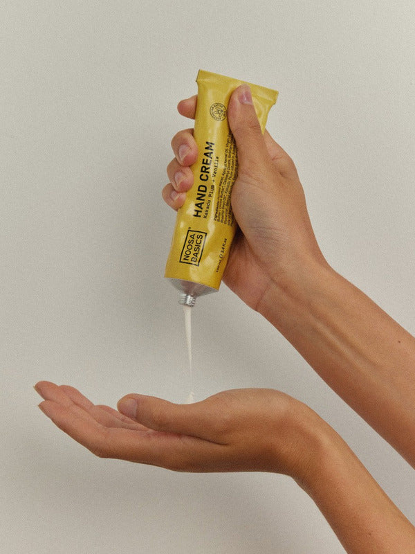 Hands squeezing a thin stream of white Noosa Basics Kakadu Plum and Vanilla Hand Cream from a yellow tube into an open palm.