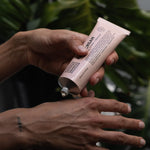 Person squeezing white Noosa Basics Kangaroo Paw Flower and Geranium Hand Cream from a pale pink tube onto the back of their hand, with green foliage behind.