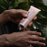 Person squeezing white Noosa Basics Kangaroo Paw Flower and Geranium Hand Cream from a pale pink tube onto the back of their hand, with green foliage behind.