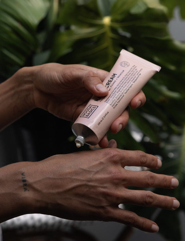 Person squeezing white Noosa Basics Kangaroo Paw Flower and Geranium Hand Cream from a pale pink tube onto the back of their hand, with green foliage behind.