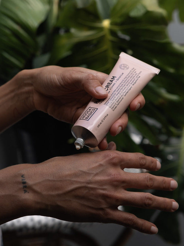 Person squeezing white Noosa Basics Kangaroo Paw Flower and Geranium Hand Cream from a pale pink tube onto the back of their hand, with green foliage behind.