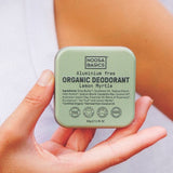 Person in a white sleeveless top holding a pale green Noosa Basics Lemon Myrtle Deodorant Tin in both hands near their chest.