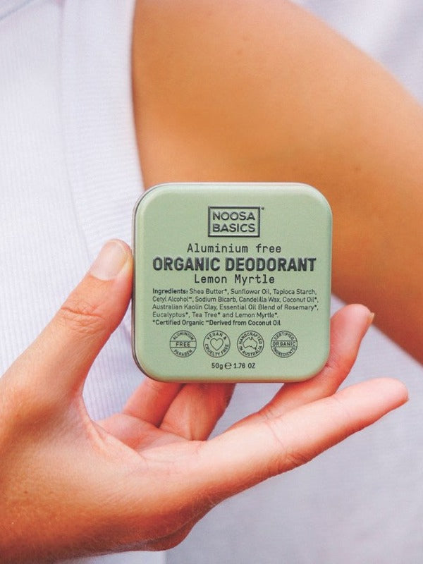 Person in a white sleeveless top holding a pale green Noosa Basics Lemon Myrtle Deodorant Tin in both hands near their chest.