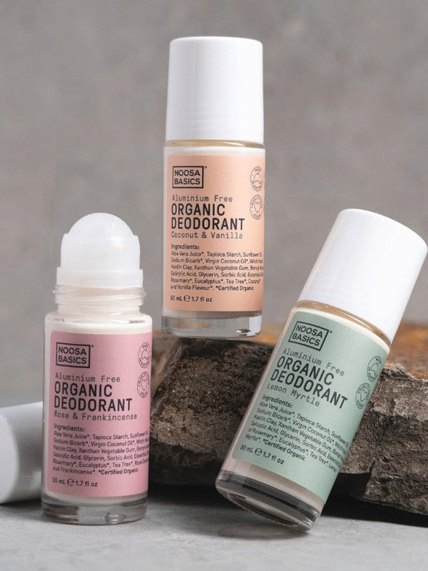 Three Noosa Basics roll-on deodorants arranged around a natural stone: Coconut and Vanilla, Lemon Myrtle, and Rose and Frankincense, with one roller ball exposed.