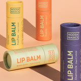 Four capped Noosa Basics lip balm tubes against peach: yellow Papaya, orange Guava and purple Chamomile tubes stand upright while a pale green Watermelon tube lies across the front.