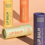 Four capped Noosa Basics lip balm tubes against peach: yellow Papaya, orange Guava and purple Chamomile tubes stand upright while a pale green Watermelon tube lies across the front.