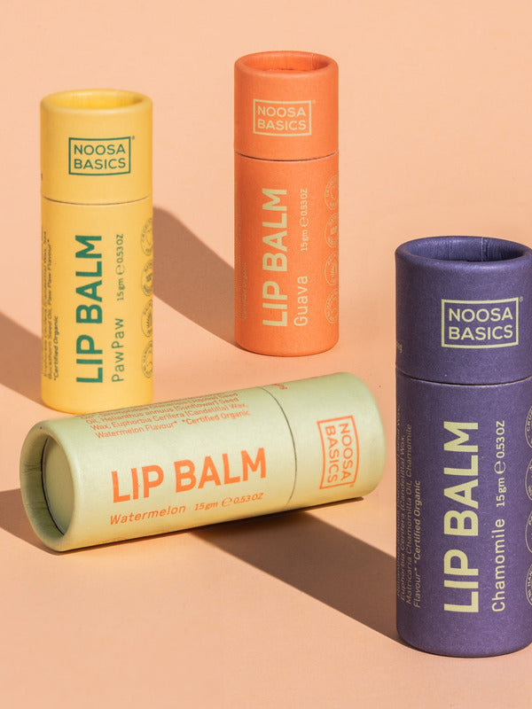 Four capped Noosa Basics lip balm tubes against peach: yellow Papaya, orange Guava and purple Chamomile tubes stand upright while a pale green Watermelon tube lies across the front.