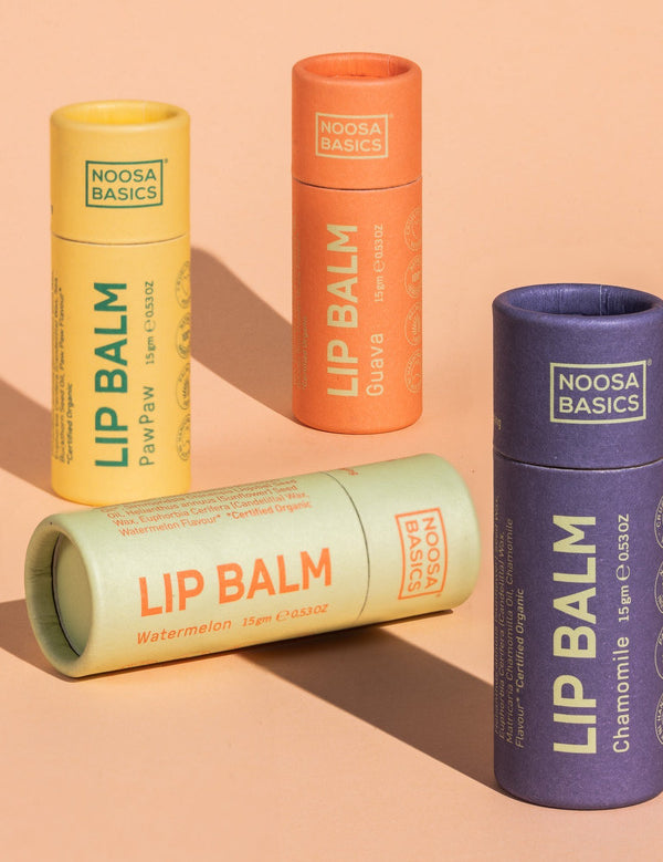 Four capped Noosa Basics lip balm tubes against peach: yellow Papaya, orange Guava and purple Chamomile tubes stand upright while a pale green Watermelon tube lies across the front.