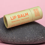 Capped pale green Noosa Basics Watermelon Lip Balm tube with orange lettering lying diagonally across a smooth dark stone against a pink background.