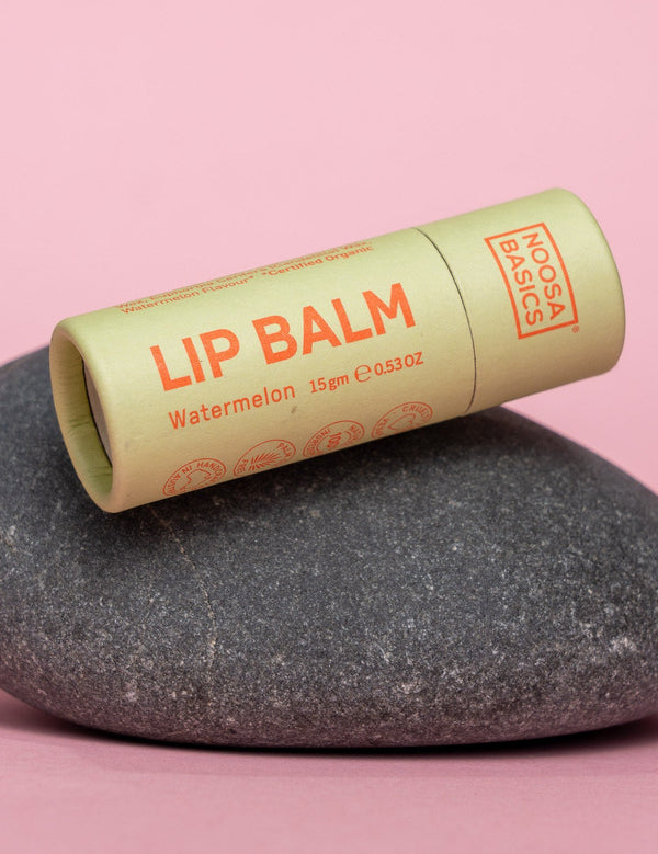 Capped pale green Noosa Basics Watermelon Lip Balm tube with orange lettering lying diagonally across a smooth dark stone against a pink background.