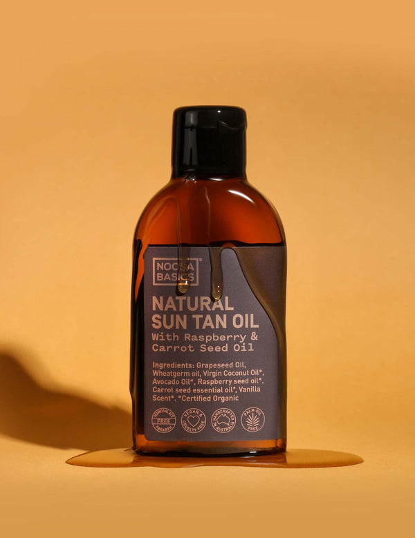 Amber Noosa Basics Natural Sun Tan Oil bottle with a black flip-top cap standing on an orange surface, surrounded by a small puddle of oil and casting a long shadow.