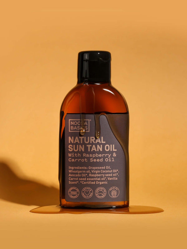 Amber Noosa Basics Natural Sun Tan Oil bottle with a black flip-top cap standing on an orange surface, surrounded by a small puddle of oil and casting a long shadow.