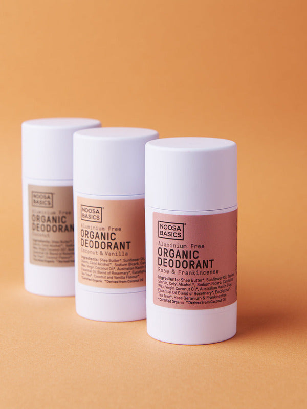 Three Noosa Basics deodorant sticks with beige, peach and pink labels arranged in a staggered row against orange.