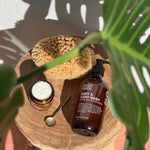 Noosa Basics Rose Geranium and Frankincense Body and Hand Wash bottle on a wooden stool with a sponge, open jar and spoon beneath green leaves.