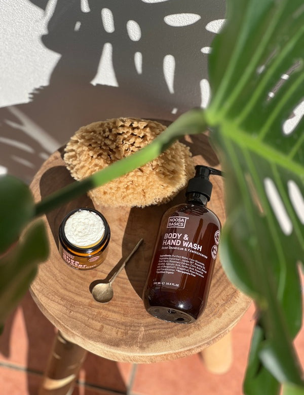 Noosa Basics Rose Geranium and Frankincense Body and Hand Wash bottle on a wooden stool with a sponge, open jar and spoon beneath green leaves.