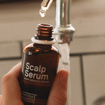 Hand holding an open amber Noosa Basics Scalp Serum bottle near a bathroom tap, with a drop forming at the tip of a glass pipette above it.