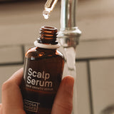 Hand holding an open amber Noosa Basics Scalp Serum bottle near a bathroom tap, with a drop forming at the tip of a glass pipette above it.