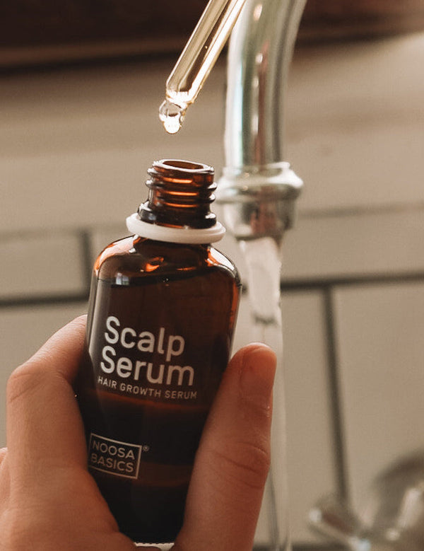 Hand holding an open amber Noosa Basics Scalp Serum bottle near a bathroom tap, with a drop forming at the tip of a glass pipette above it.