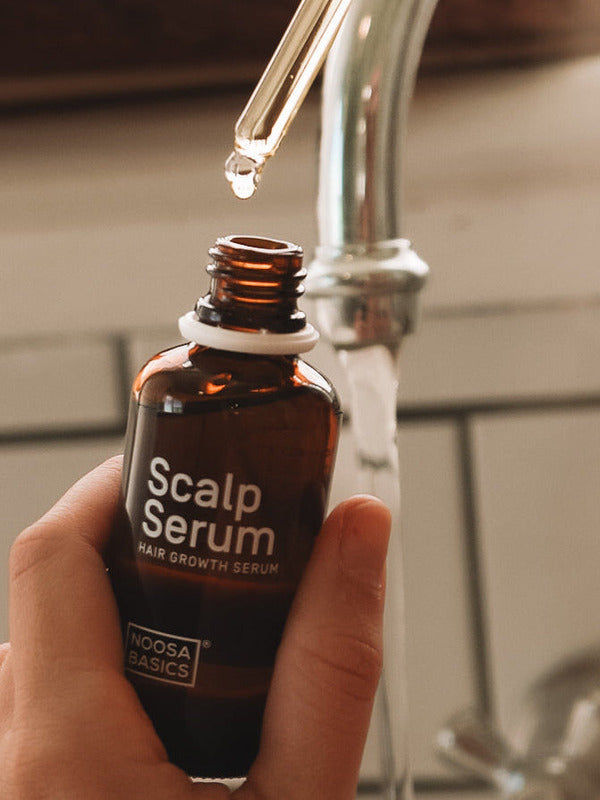 Hand holding an open amber Noosa Basics Scalp Serum bottle near a bathroom tap, with a drop forming at the tip of a glass pipette above it.