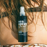 Noosa Basics Sea Spray bottle resting against a person's bare back beneath wet hair, just above patterned swimwear.