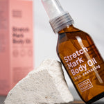 Amber Noosa Basics Stretch Mark Body Oil pump bottle leaning against a pale stone, with a pink carton standing behind it.