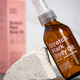 Amber Noosa Basics Stretch Mark Body Oil pump bottle leaning against a pale stone, with a pink carton standing behind it.