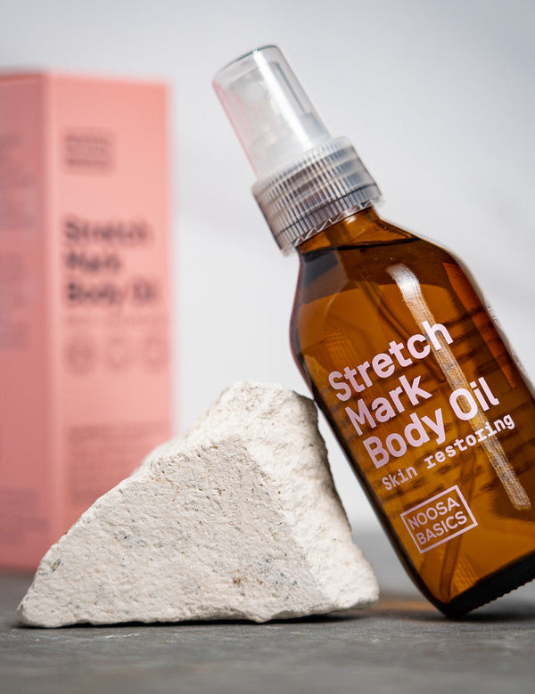 Amber Noosa Basics Stretch Mark Body Oil pump bottle leaning against a pale stone, with a pink carton standing behind it.