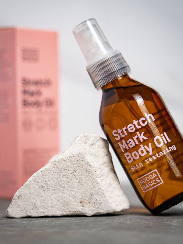 Amber Noosa Basics Stretch Mark Body Oil pump bottle leaning against a pale stone, with a pink carton standing behind it.
