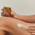 Person holding an open Noosa Basics Shea Butter jar while spreading thick white cream across the skin above a bent knee.