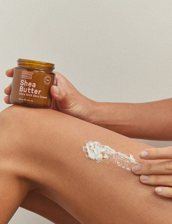 Person holding an open Noosa Basics Shea Butter jar while spreading thick white cream across the skin above a bent knee.