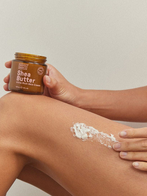 Person holding an open Noosa Basics Shea Butter jar while spreading thick white cream across the skin above a bent knee.