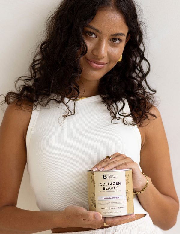 Woman in a white sleeveless top holding a jar of Nutra Naturals Collagen Beauty Vanilla at waist height against a neutral studio background.