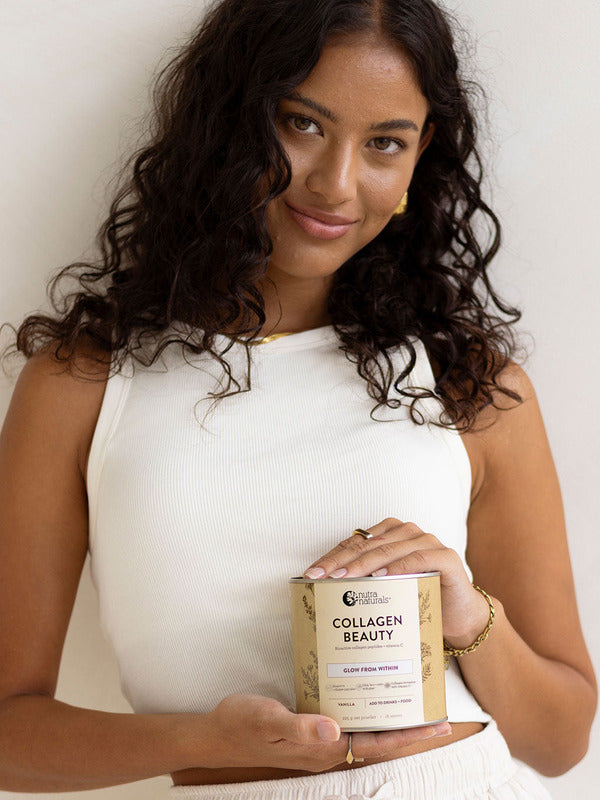Woman in a white sleeveless top holding a jar of Nutra Naturals Collagen Beauty Vanilla at waist height against a neutral studio background.