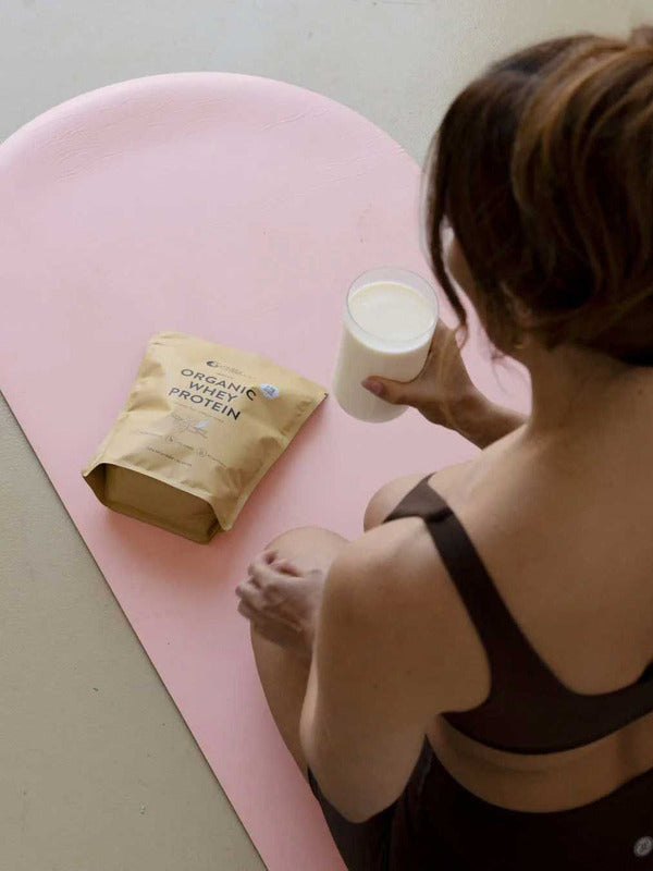 A person in workout clothes sits on a pink mat, holding a glass of milk beside a beige pouch labeled Nutra Naturals Organic Whey Protein Vanilla 750g.