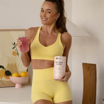 Woman in yellow activewear holding Nutra Naturals Protein Water Berry Bliss and a pink prepared drink in a bright kitchen.