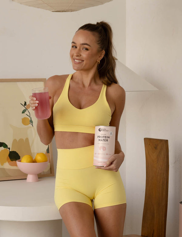 Woman in yellow activewear holding Nutra Naturals Protein Water Berry Bliss and a pink prepared drink in a bright kitchen.