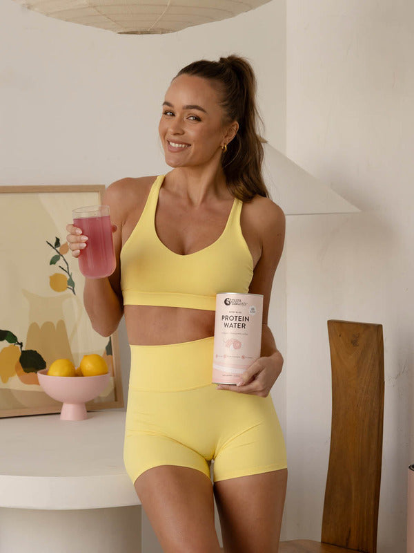 Woman in yellow activewear holding Nutra Naturals Protein Water Berry Bliss and a pink prepared drink in a bright kitchen.