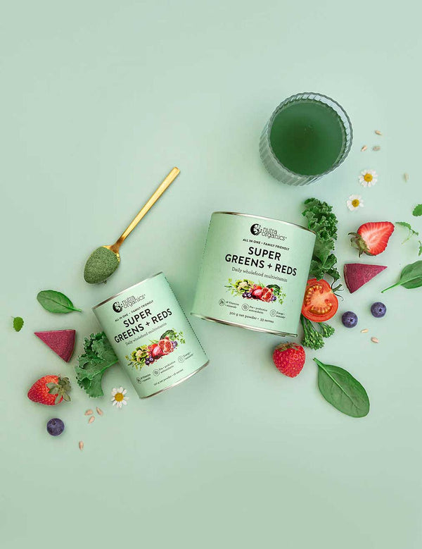Two containers of Nutra Naturals Super Greens + Reds with a scoop, surrounded by superfoods, leafy greens, strawberries, spinach, blueberries, tomato, and a green drink in a glass, all set on a pale green background.