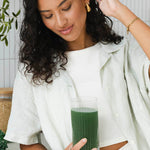 Woman looking down at a tall glass of dark green drink held in one hand, with her other hand raised to her hair.
