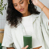 Woman looking down at a tall glass of dark green drink held in one hand, with her other hand raised to her hair.