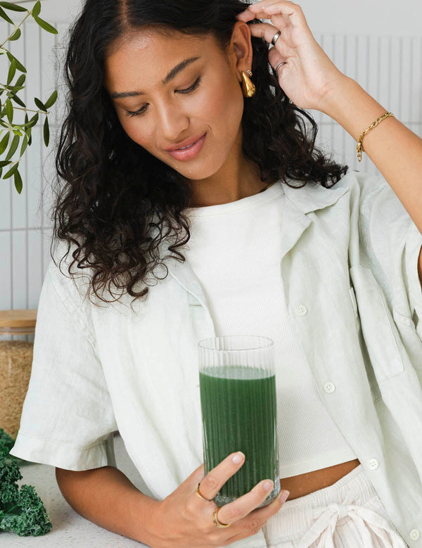 Woman looking down at a tall glass of dark green drink held in one hand, with her other hand raised to her hair.