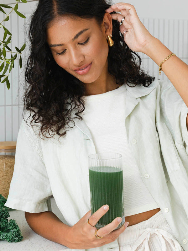 Woman looking down at a tall glass of dark green drink held in one hand, with her other hand raised to her hair.