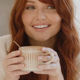 Lifestyle portrait of a smiling red-haired woman holding a warm caramel-coloured drink in a ceramic mug for Nutra Organics Collagen Beauty Caramel.