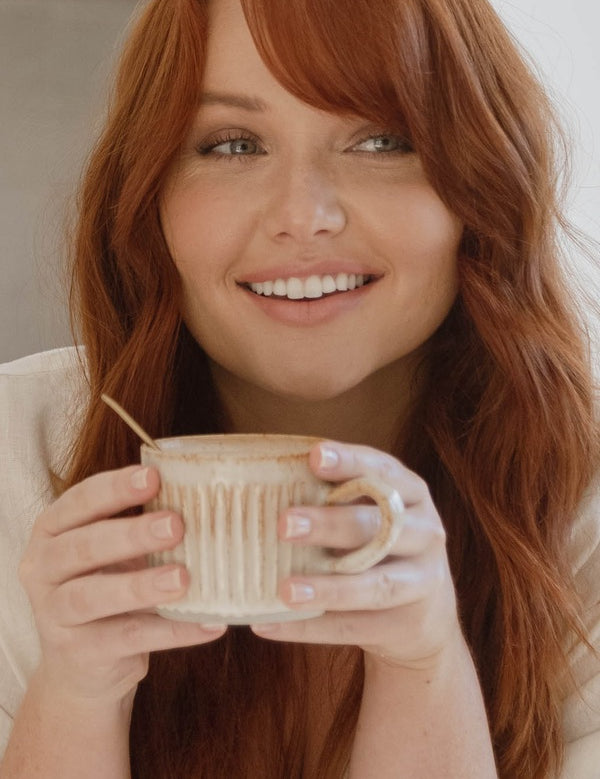 Lifestyle portrait of a smiling red-haired woman holding a warm caramel-coloured drink in a ceramic mug for Nutra Organics Collagen Beauty Caramel.