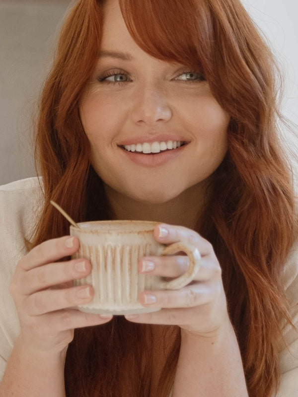 Lifestyle portrait of a smiling red-haired woman holding a warm caramel-coloured drink in a ceramic mug for Nutra Organics Collagen Beauty Caramel.