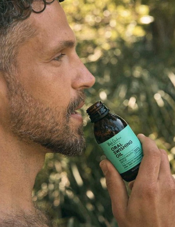 Man outdoors holding an open Oral Swishing Oil bottle close to his lips, with sunlit foliage behind him.
