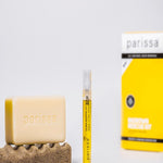 Pale rectangular Parissa bar resting on a textured brown pad beside a slim yellow-labelled tube, with the yellow Ingrown Rescue Kit box in the background.
